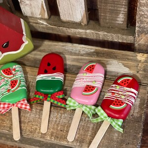 May include: Four wooden watermelon popsicle decorations with green, red, and pink paint. Each popsicle has a wooden stick and a ribbon bow.
