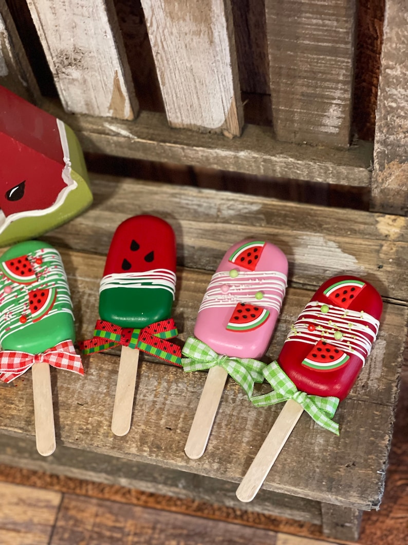 May include: Four wooden watermelon popsicle decorations with green, red, and pink paint. Each popsicle has a wooden stick and a ribbon bow.