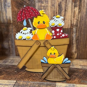 May include: Wooden duck-themed decorative set. Features a large basket with a yellow duck, daisies, a red polka-dot umbrella, and a red polka-dot boot. A smaller basket holds a duck in water. The set is painted in bright colors.