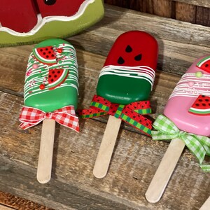 May include: Three watermelon popsicle-shaped decorations with wooden sticks and red and white gingham bows. The popsicles are painted to look like watermelon with green, red, and black details.