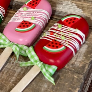 May include: Two watermelon-shaped cake pops on wooden sticks with green and white gingham ribbon bows. The cake pops are decorated with white frosting, green and white sprinkles, and a red watermelon slice with black seeds.