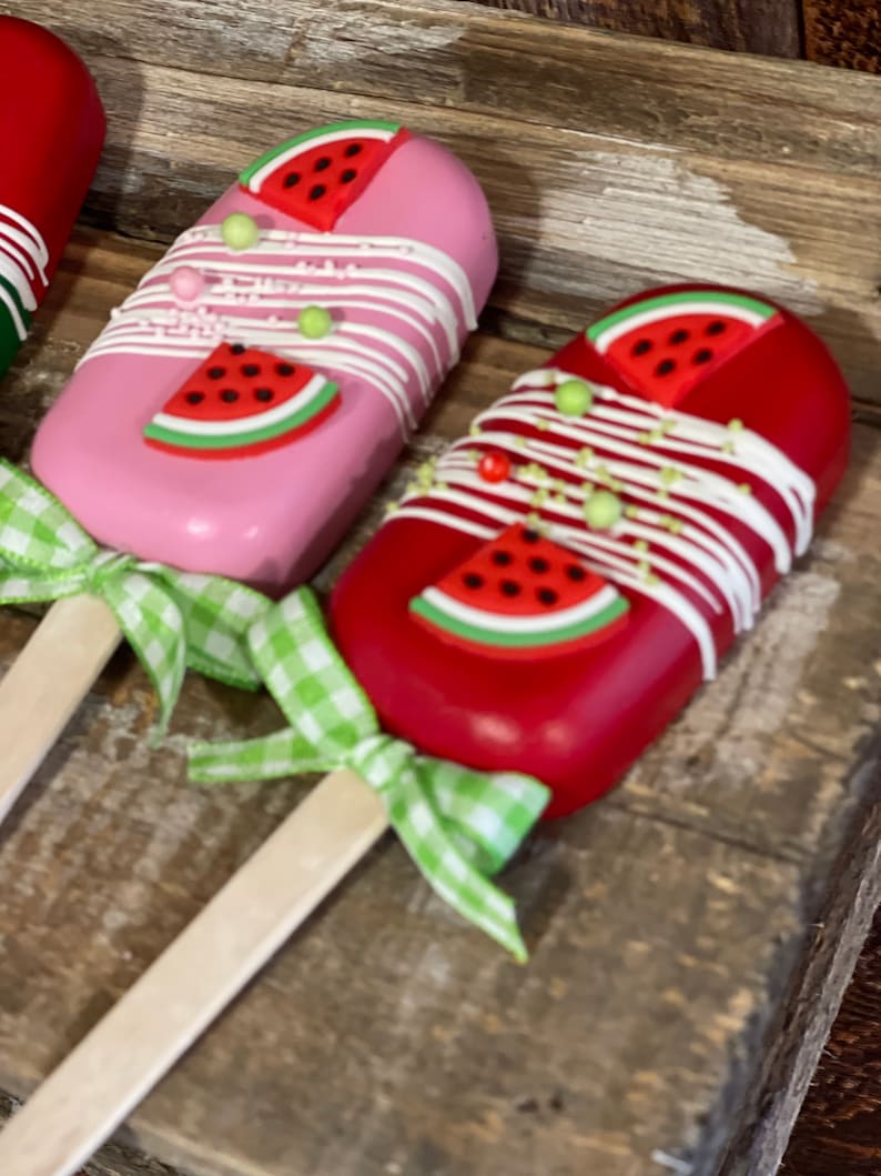 May include: Two watermelon-shaped cake pops on wooden sticks with green and white gingham ribbon bows. The cake pops are decorated with white frosting, green and white sprinkles, and a red watermelon slice with black seeds.
