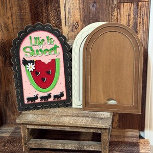 May include: Wooden decorative sign with a watermelon design and the text "Life is Sweet". The sign has a black scalloped border and a pink background. Two additional wooden arched frames are also visible.