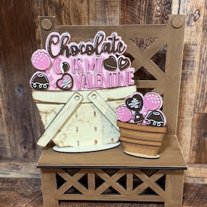 May include: Wooden craft with a pink and brown color scheme. The craft features a sign that says "Chocolate is my Valentine" and two baskets filled with chocolate candies.