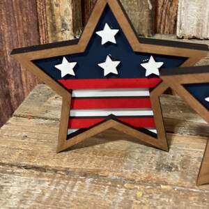 May include: A wooden star-shaped decoration with a red, white and blue American flag design. The star is painted with a dark blue background and white stars. The stripes are red and white.