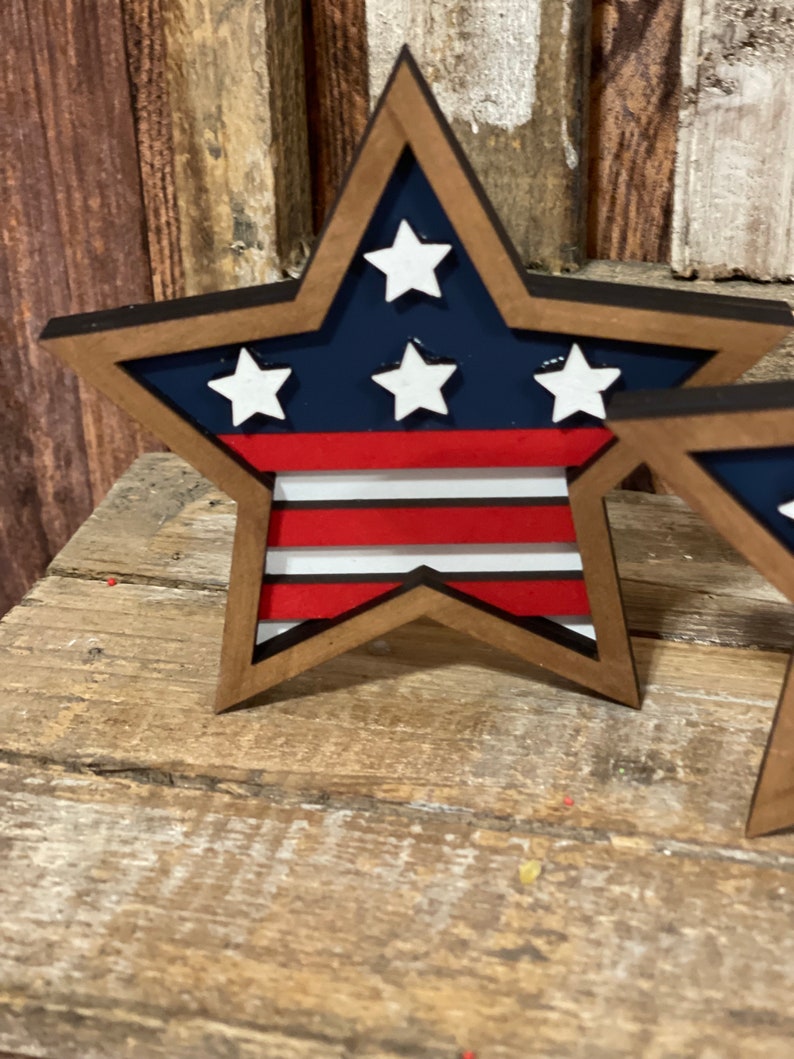 May include: A wooden star-shaped decoration with a red, white and blue American flag design. The star is painted with a dark blue background and white stars. The stripes are red and white.
