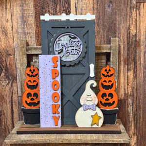 May include: A decorative Halloween display featuring a dark gray wooden door with a circular sign reading It's Freaking Bats in here, a light purple sign with the word SPOOKY, two stacks of orange jack-o'-lanterns, and a white ghost with a star.