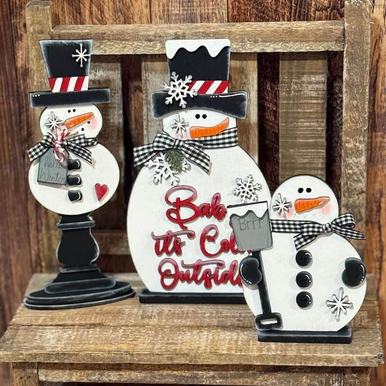 May include: Three wooden snowman decorations with black hats and scarves. The middle snowman is the largest and has the text "Babe it's Cold Outside" in red letters. The smallest snowman is holding a white cup with the text "Brrr" on it.