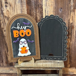 May include: Halloween-themed wooden sign with a ghost holding a pumpkin, the words "hey BOO", and purple bats. The sign is brown with a black chalkboard background. A second black scalloped-edge sign is next to it.