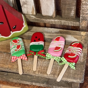 May include: Four watermelon popsicle-shaped cake pops with green, pink, and red frosting and white chocolate accents. Each cake pop has a wooden stick and a red and white gingham ribbon.