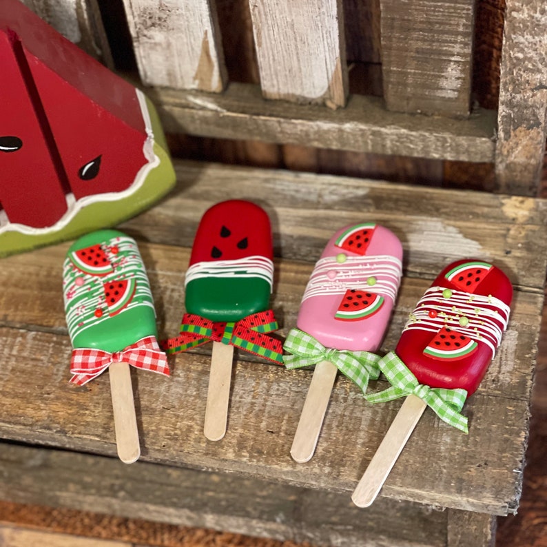 May include: Four watermelon popsicle-shaped cake pops with green, pink, and red frosting and white chocolate accents. Each cake pop has a wooden stick and a red and white gingham ribbon.