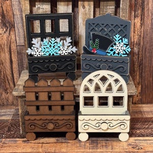 May include: Four decorative wooden window box displays. One black window with blue and white snowflakes, one dark gray lattice with a top hat and snowflakes, one brown picket fence, and one cream arched window. Each features a scrollwork base.
