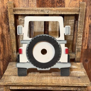 May include: A flat, off-white wooden cutout shaped like the rear view of a vehicle. It features a prominent black spare tire mounted in the center, grey bumpers, small red taillights, and two grey side mirrors extending from the upper frame.