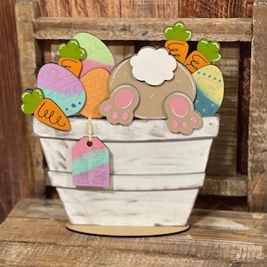 May include: A white wooden planter with a distressed finish, featuring a bunny's backside, colorful Easter eggs, and carrots. A tag with the words "Easter Egg Hunt" hangs from the planter. The Easter-themed decoration is set on a wooden surface.