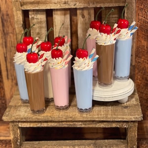 May include: A set of six miniature milkshakes with whipped cream and cherries on top. The milkshakes are in clear glass cups with colorful straws. The milkshakes are arranged on a wooden shelf.