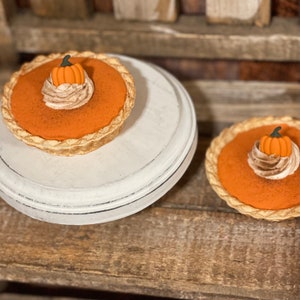 May include: Two miniature pumpkin pies. The pies have a bright orange filling, a whipped cream topping, and a small pumpkin decoration. One pie sits on a white, distressed-look cake stand. The other pie is on a wooden surface.