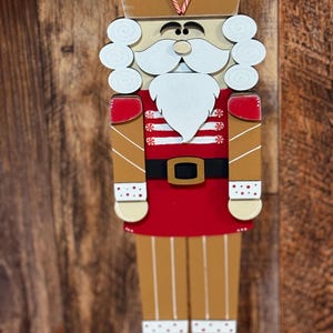 Peut inclure: Une figurine de casse-noisette en bois avec un uniforme ray&eacute; rouge et blanc, une couronne dor&eacute;e avec des cannes en sucre et une barbe blanche. Le casse-noisette est debout sur une surface en bois.