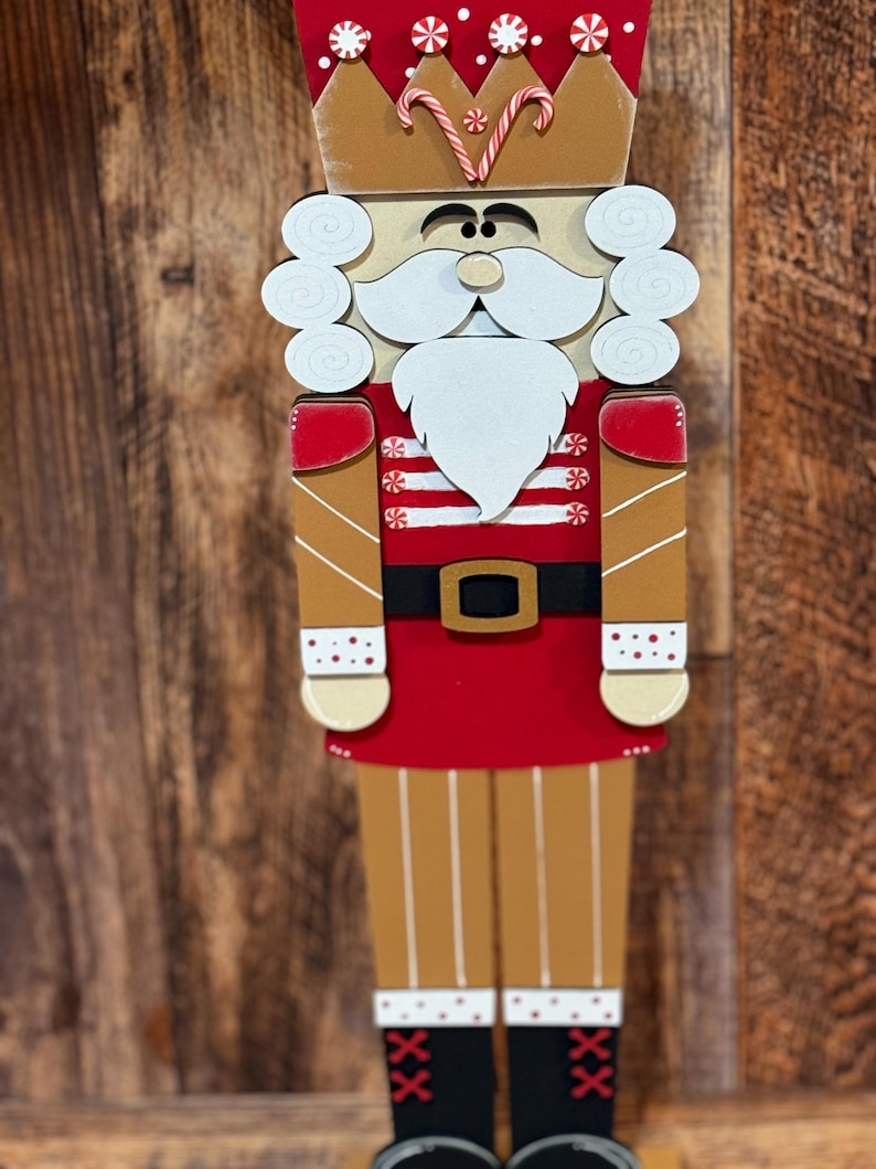 Peut inclure: Une figurine de casse-noisette en bois avec un uniforme ray&eacute; rouge et blanc, une couronne dor&eacute;e avec des cannes en sucre et une barbe blanche. Le casse-noisette est debout sur une surface en bois.