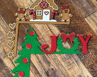 May include: A Christmas-themed wooden sign with the word "JOY" in red and green letters. The sign also features a gingerbread house, two gingerbread people, a green Christmas tree, and a red bow.