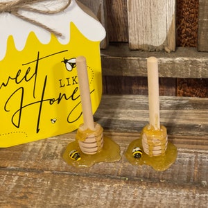 May include: Two wooden honey dippers with dripping honey and small yellow and black bees on a wooden surface. The dippers are on a yellow background with the text "Sweet Like Honey".