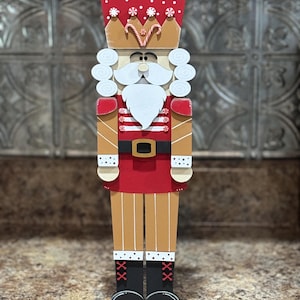 Peut inclure: Une figurine de casse-noisette en bois avec un chapeau rouge et blanc, une barbe blanche et un uniforme ray&eacute; rouge et blanc. Le casse-noisette est debout sur une base en bois.