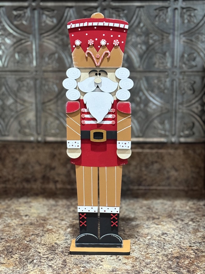 Peut inclure: Une figurine de casse-noisette en bois avec un chapeau rouge et blanc, une barbe blanche et un uniforme ray&eacute; rouge et blanc. Le casse-noisette est debout sur une base en bois.