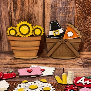 May include: Wooden craft decorations for fall and Thanksgiving. The decorations include a basket with a pilgrim hat and a pie, a basket with sunflowers, a red apple, a white cupcake, a ruler, a pencil, a daisy flower, and a letter with a heart.
