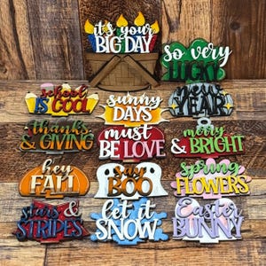 May include: A collection of 14 wooden signs with various holiday and seasonal themes. The signs are painted in bright colors and feature phrases like "It's your Big Day", "So Very Lucky", "School is Cool", "Sunny Days", "New Year", "Thanks & Giving", "Must Be Love", "Merry & Bright", "Hey Fall", "Say Boo", "Spring Flowers", "Stars & Stripes", "Let it Snow", and "Easter Bunny".