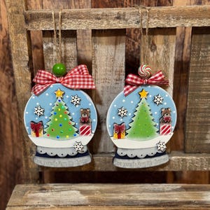 May include: Two Christmas ornaments shaped like snow globes. Each ornament features a green Christmas tree, a teddy bear, and a wrapped gift. The ornaments are decorated with snowflakes and topped with a red and white checkered bow.