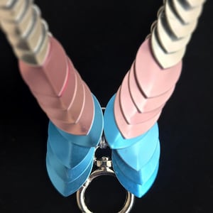 May include: A scale maille necklace featuring overlapping silver, pastel pink, and light blue metal scales arranged in a V-shape pattern. The necklace terminates at the bottom with a circular silver metal lobster clasp ring connector.