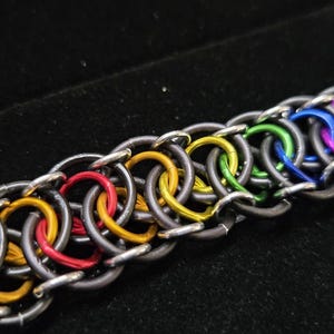Pode incluir: Uma pulseira de cota de malha feita de anéis de metal entrelaçados. O design apresenta um padrão repetitivo de anéis cinza-escuros combinados com anéis de cores vibrantes em tons de vermelho, amarelo, verde, azul e roxo, criando um efeito arco-íris.