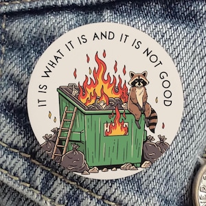 May include: A round, white button with a cartoon illustration of a dumpster fire. A raccoon sits on the dumpster, with the text "IT IS WHAT IT IS AND IT IS NOT GOOD" around the edge. The dumpster is green, and the fire is orange and yellow.
