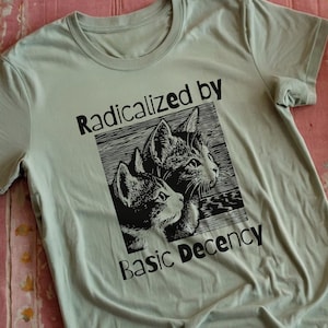 May include: A light green t-shirt with a black and white graphic of two cats and the text "Radicalized by Basic Decency." The shirt is made of soft material and has short sleeves.
