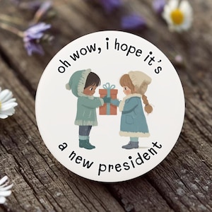 May include: A round, white sticker with the text "oh wow, I hope it's a new president." The sticker features an illustration of two children exchanging a gift. The children are wearing winter coats and hats. The background is a wooden surface with flowers.