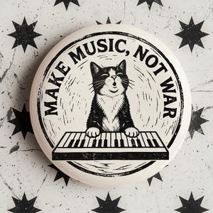 May include: A round white pinback button featuring a black and white illustration of a cat playing a piano. The text "MAKE MUSIC, NOT WAR" is printed in a circular arc around the cat. The design has a distressed, ink-stamped aesthetic.