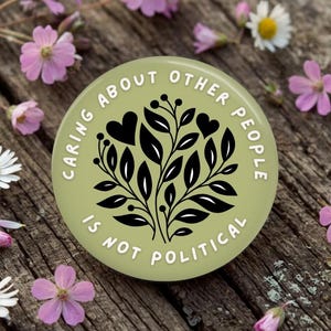 May include: A round, light green button with the text "CARING ABOUT OTHER PEOPLE IS NOT POLITICAL" in white. The button features a black floral design with hearts. The button is surrounded by pink flowers and daisies on a wooden surface.