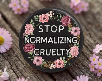 May include: A black circular button with the text "STOP NORMALIZING CRUELTY" surrounded by pink and burgundy flowers and green leaves. The button is set on a weathered wooden surface, with scattered pink flowers.