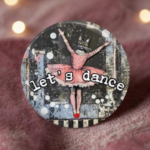 May include: A circular pin featuring a black cat with yellow eyes wearing a pink tutu and red ballet slippers. The background is a dark, textured collage with white polka dots and the text let's dance written in a white, bold, outlined font.