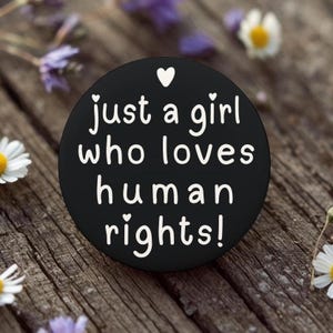 May include: A black round pop socket with the text "just a girl who loves human rights!" in white. The design includes small white hearts. The pop socket is on a wooden surface with flowers.