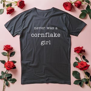 May include: A dark gray t-shirt with the text "never was a cornflake girl" in white. The shirt is surrounded by red roses on a pink background. The t-shirt is a casual wear item.