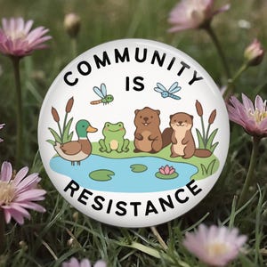 May include: A round button with the text "COMMUNITY IS RESISTANCE" in black. The button features a cartoon illustration of a duck, frog, beavers, and otters around a pond with dragonflies and cattails.
