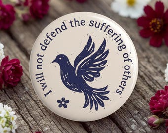 May include: A circular pinback button with a cream-colored background featuring a dark blue graphic of a flying bird. The text "I will not defend the suffering of others" is printed in a dark blue circular arc around the bird, with a small flower icon.