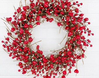 May include: A circular wreath composed of red heart-shaped berries and small white berries on brown twigs. The wreath is set against a white brick wall, ideal for Valentine's Day or other celebrations.
