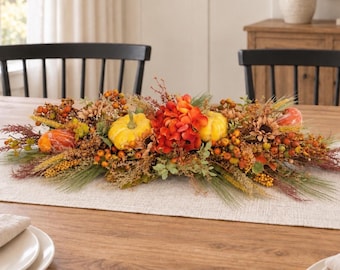 May include: A seasonal table centrepiece featuring artificial yellow pumpkins, vibrant orange hydrangea blossoms, pine needles, dried grasses, and clusters of small berries in shades of orange, yellow, and brown, arranged in a long, horizontal display.