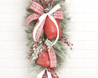 May include: A vertical Christmas decoration with two red glitter heart ornaments, red and white plaid bows, and white ribbons. It features faux pine needles, red berries, and white frosted accents. The arrangement is set against a white brick wall background.