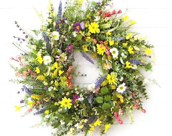 May include: A circular wreath composed of a dense arrangement of artificial wildflowers and greenery. The display features vibrant yellow, purple, pink, and white blossoms interspersed with various green leaves, stems, and small berry-like clusters.
