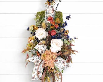 May include: A vertical floral arrangement featuring white peonies, orange hydrangeas, and blue sprigs attached to a moss-covered base. The piece is accented with a patterned ribbon featuring a floral print and long brown satin ribbons hanging down.
