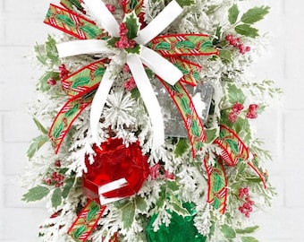 May include: A teardrop-shaped festive swag featuring flocked evergreen branches, red berries, and green holly leaves. A large white ribbon bow sits at the top, accented by a red and green patterned ribbon. Two large faceted ornaments, one red and one green, hang below.