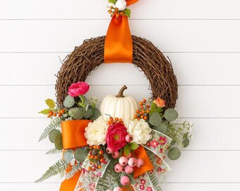 May include: A grapevine wreath featuring a central white pumpkin, accented with vibrant pink and cream flowers, orange berries, green eucalyptus leaves, and fern fronds. A bright orange satin ribbon hangs from the top and decorates the base.