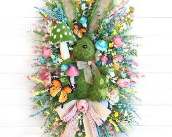 May include: An Easter bunny wreath with a moss-covered body, decorated with colourful flowers, butterflies, and mushroom accents. The wreath features long, textured ears and a burlap bow with pink ribbon.